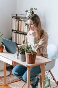 A young woman works on a laptop in a cozy home office surrounded by plants and bookshelves.